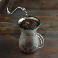 Coffee bloom during brewing in glass carafe with stainless steel filter on wooden surface